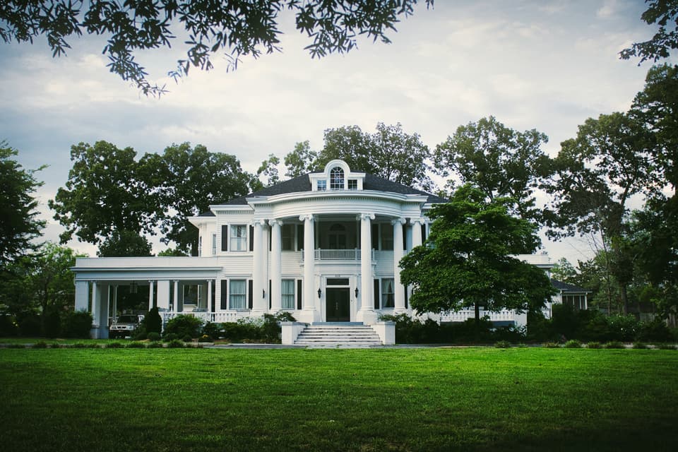 A large white house behind a wide lawn and mature shade trees